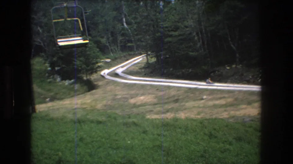1976 Mt. Bromley home-movie frame showing a yellow chairlift seat passing over a grassy slope with a winding white track bordered by dense trees.