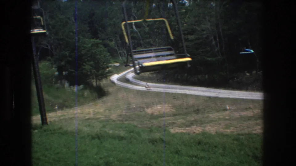 A closer 1976 chairlift view at Mt. Bromley shows a yellow seat crossing the frame above a curving white track and wooded resort slope.