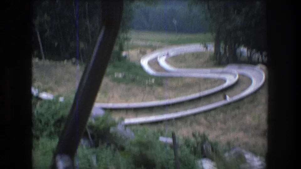 From the moving chairlift, the 8mm frame looks down toward a winding white resort track at Mt. Bromley with a small distant person visible on or near the track.