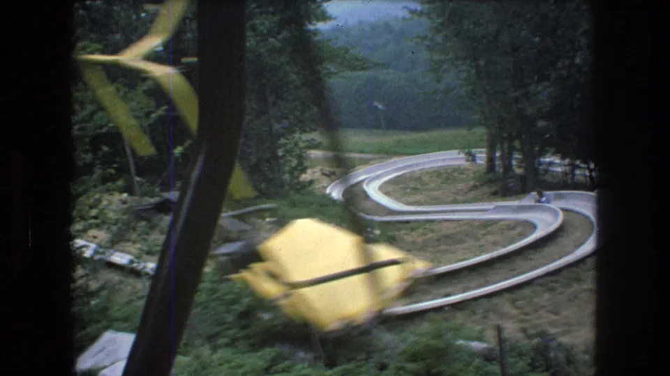 A yellow chairlift chair moves across the foreground while the Mt. Bromley alpine slide curves through trees below in this 1976 home-movie frame.