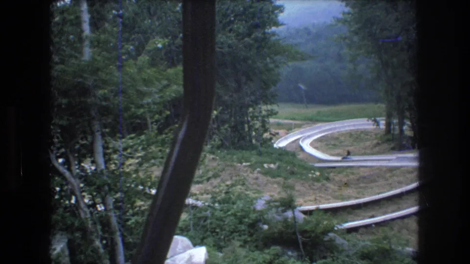 The alpine slide at Mt. Bromley curves through a wooded slope, with small distant riders visible along the track in 1976 8mm footage.