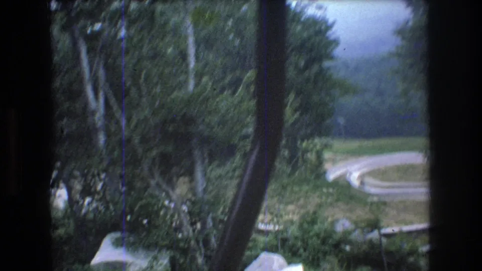 A motion-blurred 1976 chairlift view frames trees, a vertical lift support, and the pale curve of the Mt. Bromley alpine slide below.