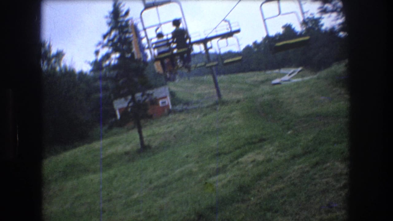 1976 Mt. Bromley home-movie frame showing chairlift seats over a green slope, with evergreen trees and a small red resort building in the background.