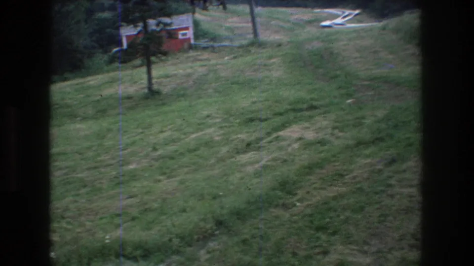 1970s 8mm frame of the Mt. Bromley hillside, showing a small tree on the slope, a red building, lift structure, and green summer grass.
