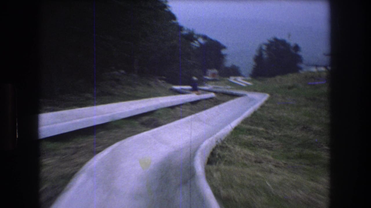 1976 8mm footage at Mt. Bromley shows a first-person view entering a curving concrete alpine slide bordered by grass and trees, with a distant rider visible ahead.