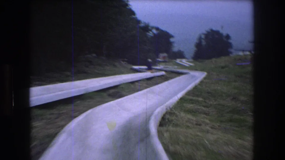 1976 8mm footage at Mt. Bromley shows a first-person view entering a curving concrete alpine slide bordered by grass and trees, with a distant rider visible ahead.