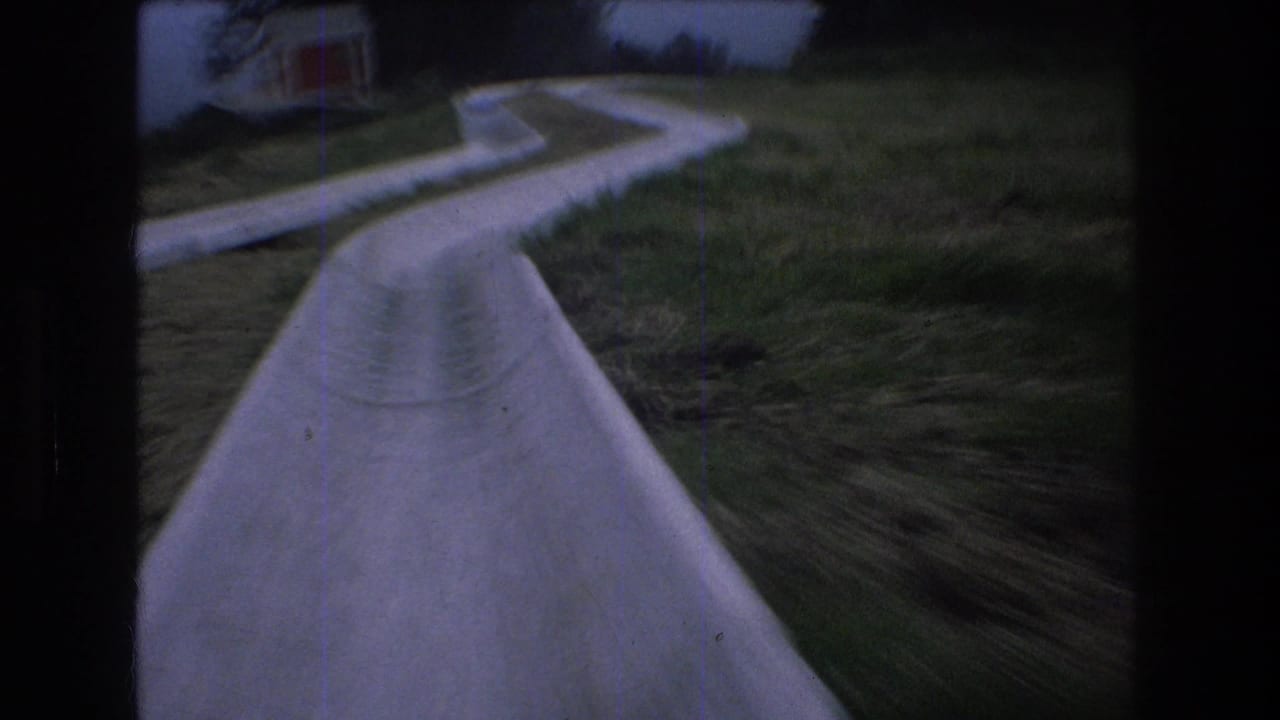 1976 Mt Bromley home-movie frame showing a rider's view down a pale concrete water slide bordered by grass, with a small building partly visible ahead.