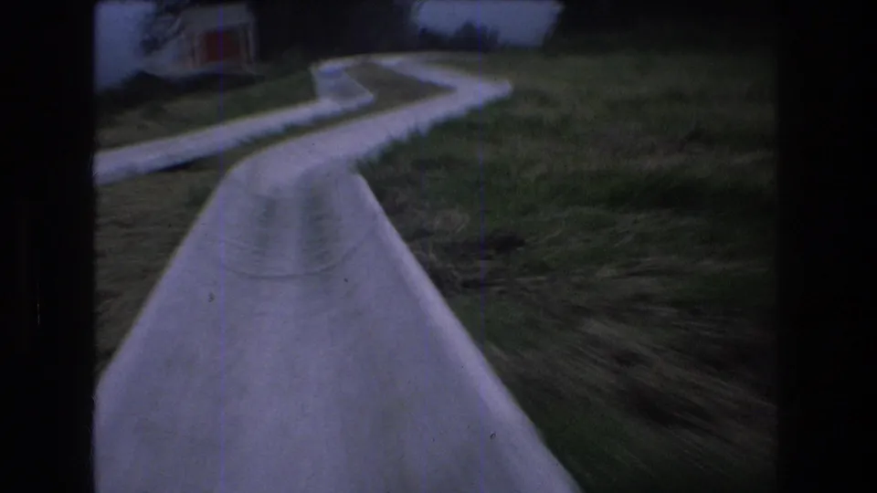 1976 Mt Bromley home-movie frame showing a rider's view down a pale concrete water slide bordered by grass, with a small building partly visible ahead.