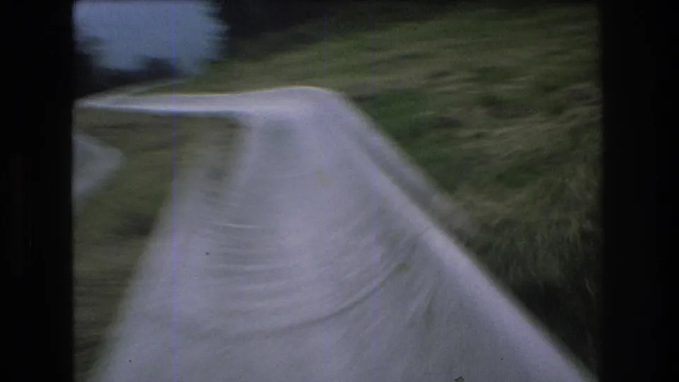 1976 Mt Bromley frame with the camera moving quickly along a concrete water slide, showing the curving channel, grass, and dark tree line at the edge of the frame.