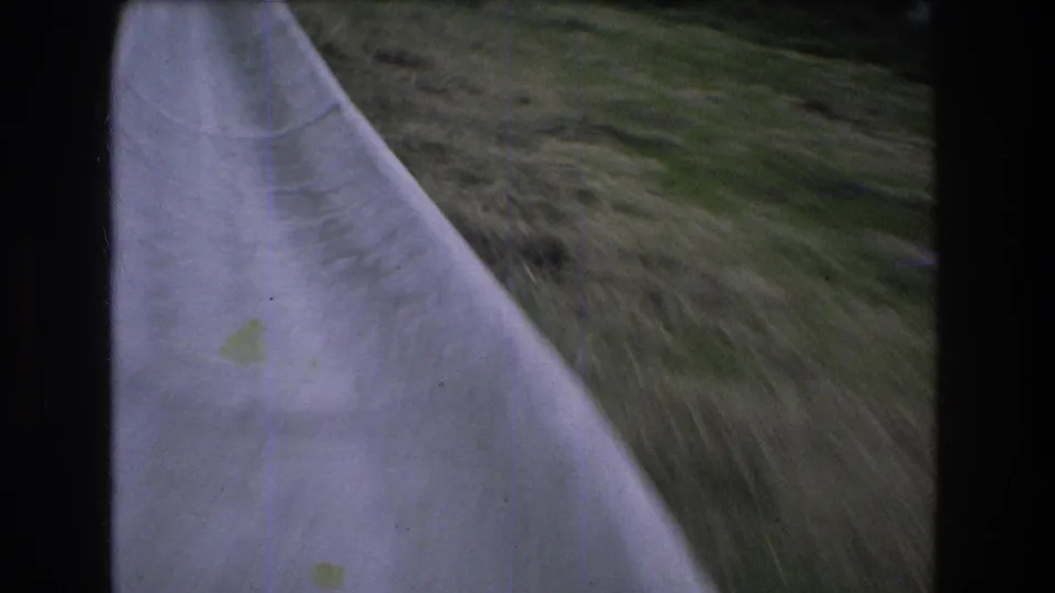 1976 Mt Bromley home-movie frame showing a close, tilted rider's view along the concrete slide wall with grass rushing past on the hillside.