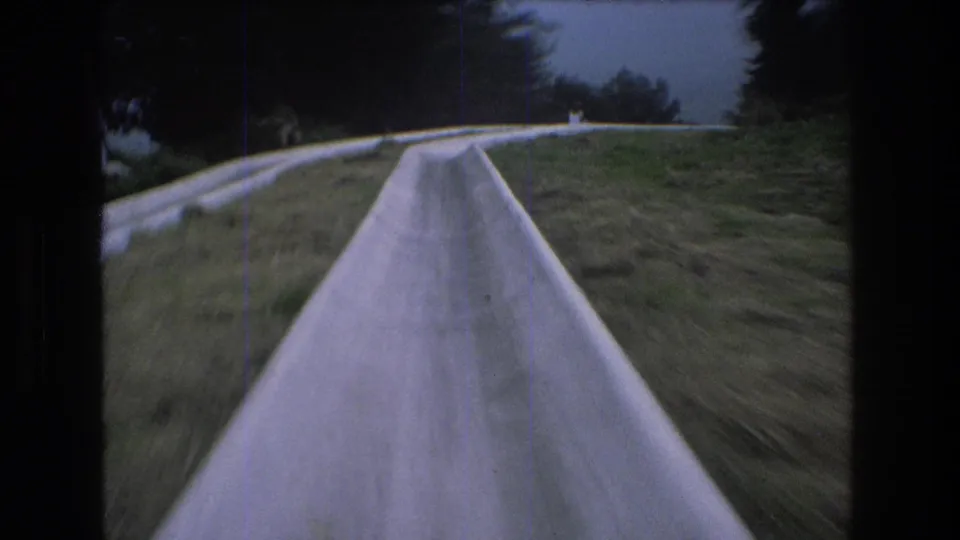 1976 Mt Bromley frame showing the slide stretching ahead through grass toward a higher section of the hillside, with trees and cloudy sky in the distance.