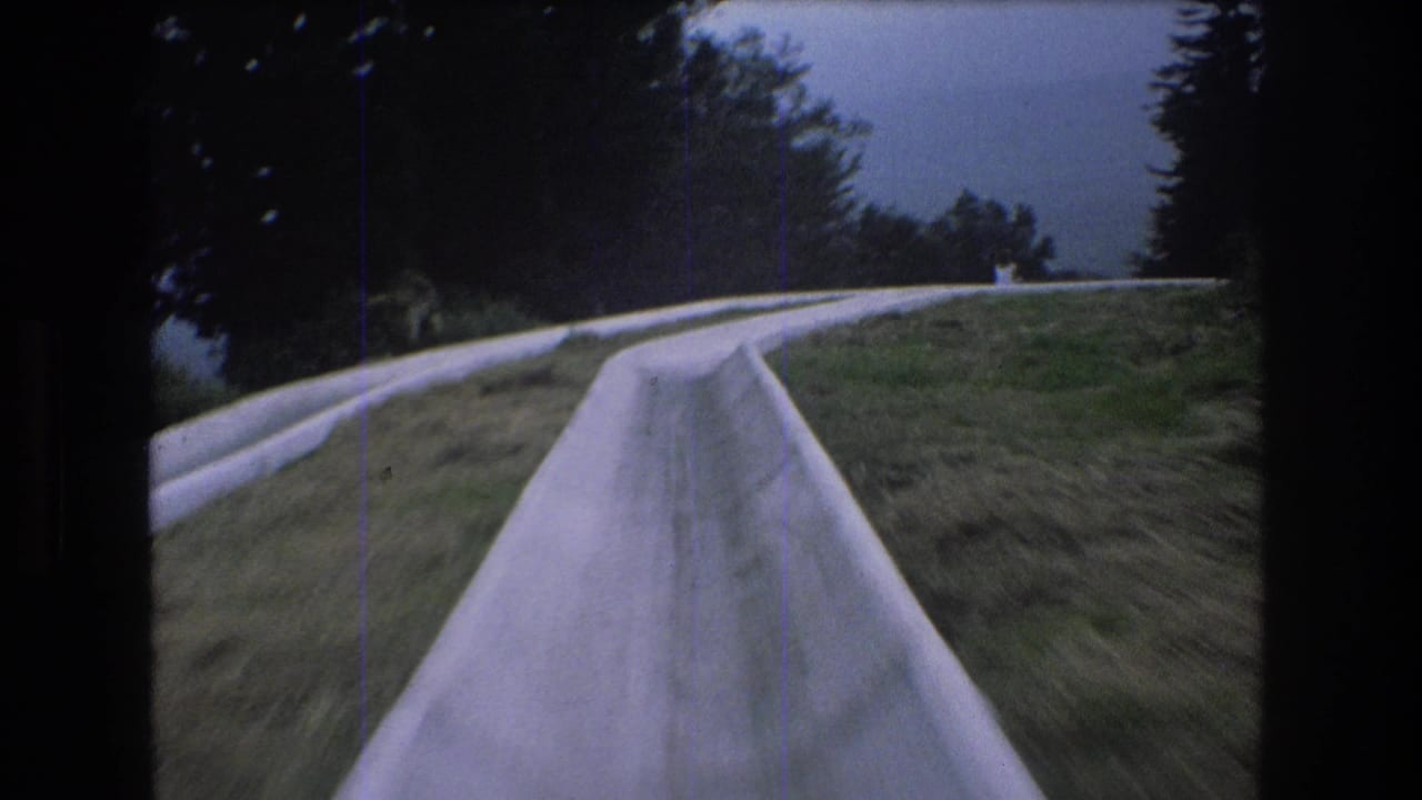 First-person 1976 8mm view at Mt Bromley, Vermont, looking down a narrow concrete luge or alpine slide chute as it curves through grass and trees.