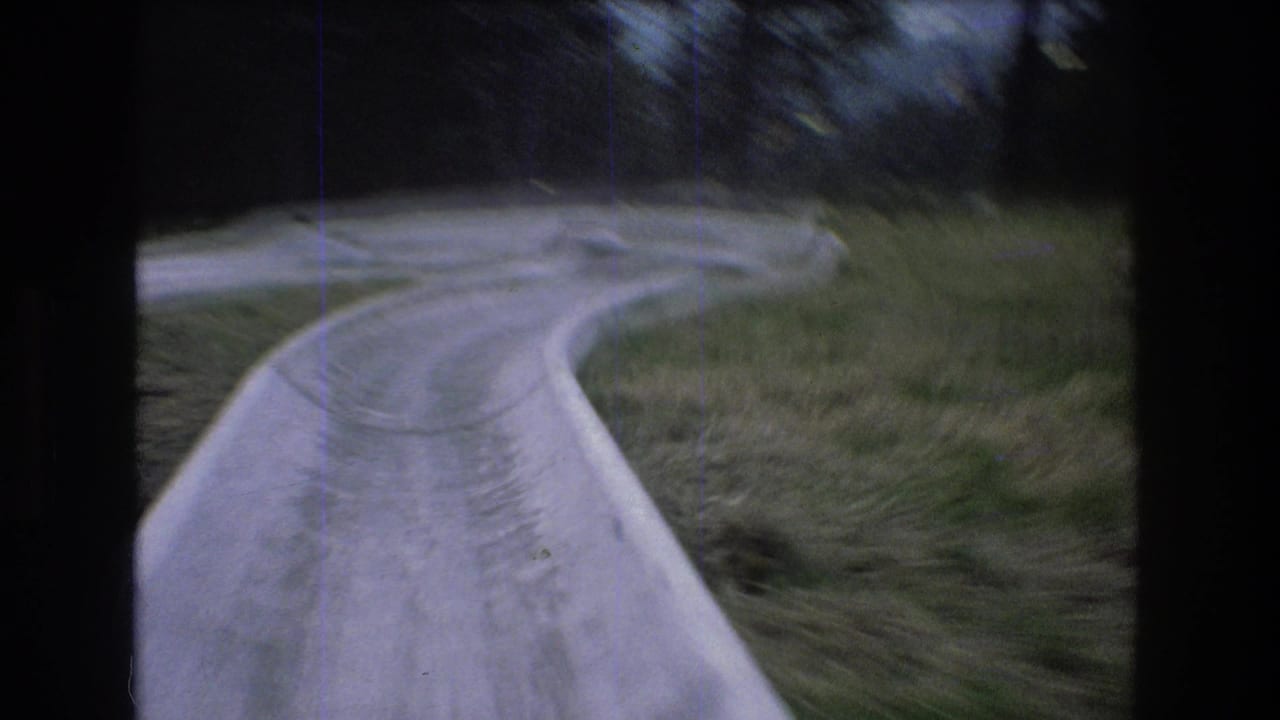 1976 Mt Bromley, Vermont 8mm frame showing a rider's view down a concrete alpine slide as it bends through a grassy hillside with dark trees beyond.