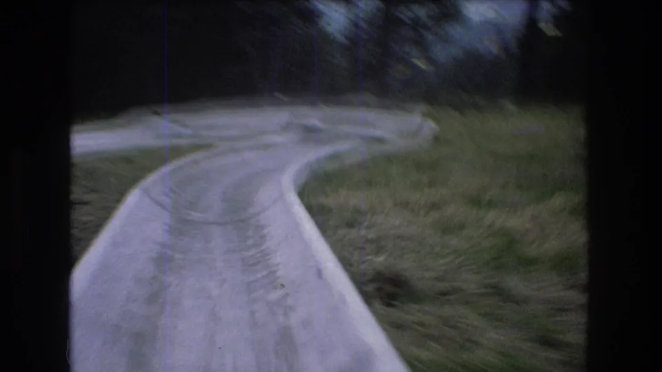 1976 Mt Bromley, Vermont 8mm frame showing a rider's view down a concrete alpine slide as it bends through a grassy hillside with dark trees beyond.
