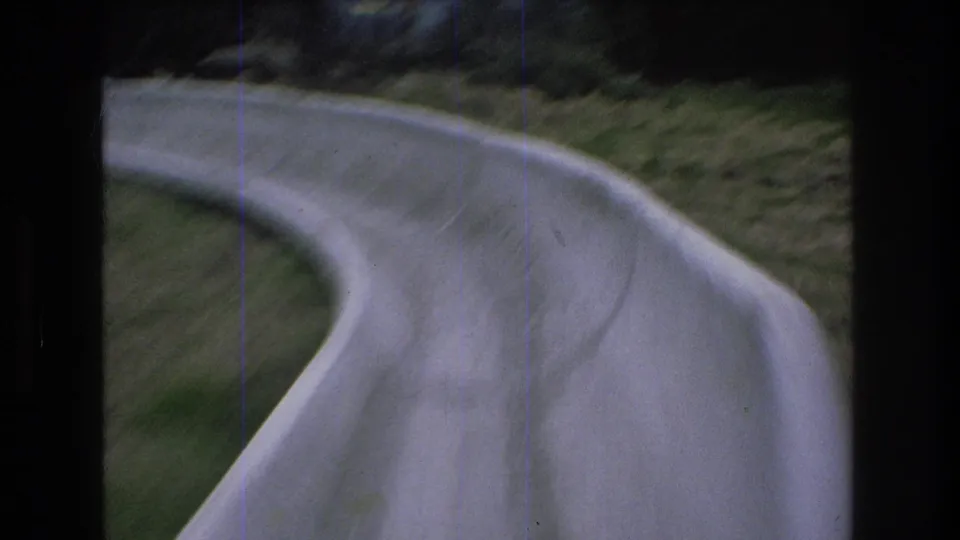1976 Mt Bromley, Vermont 8mm frame showing a close, blurred point-of-view of a banked concrete alpine slide turn bordered by grass.