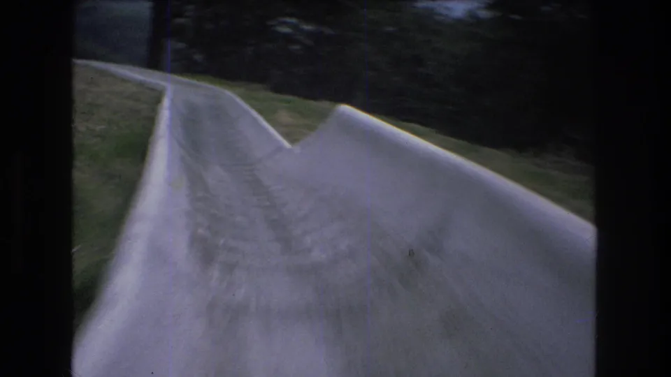 1976 8mm home-movie frame from Mt Bromley, Vermont, showing a point-of-view look down a pale concrete slide with raised sides, grass, and trees ahead.