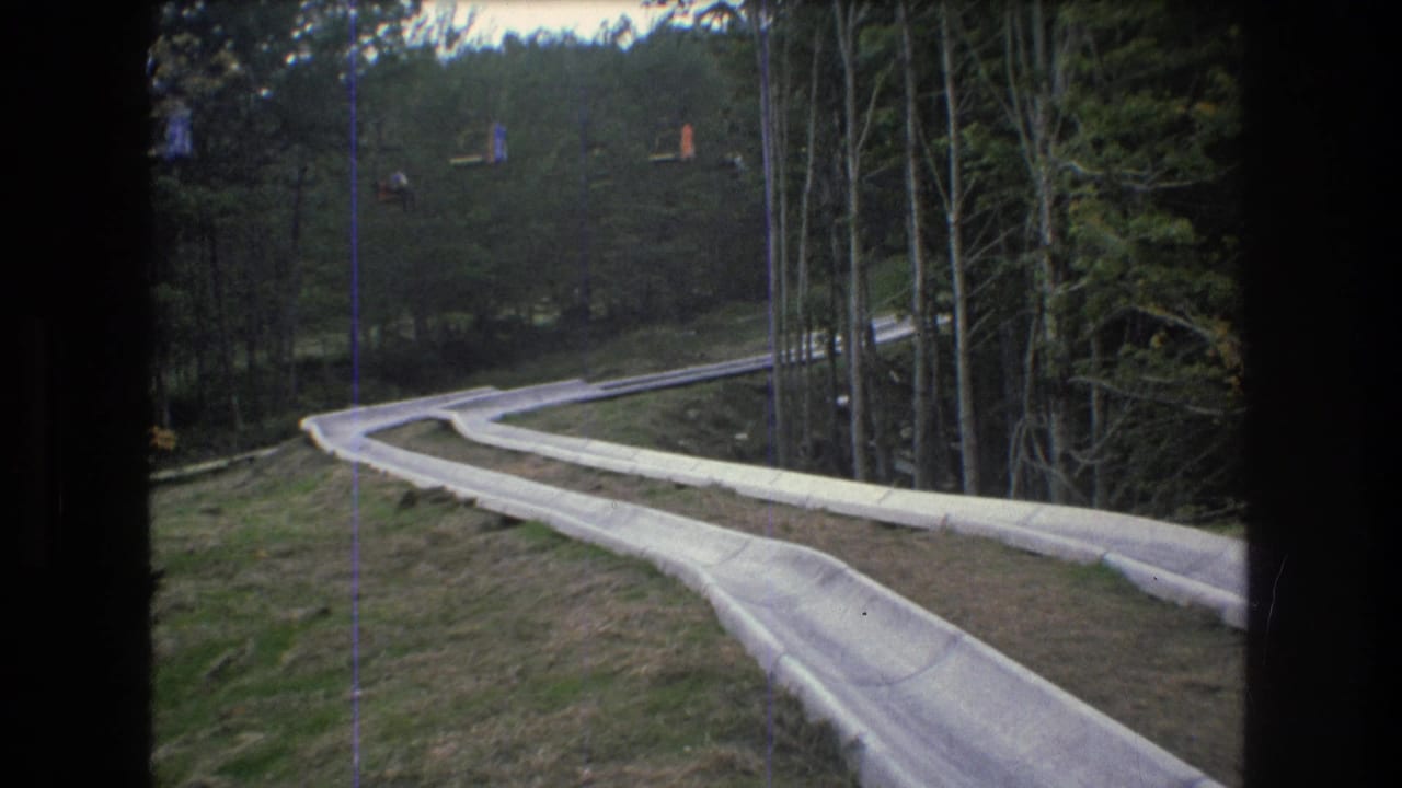 1976 Mt Bromley, Vermont 8mm frame showing a concrete water slide curving down a grassy forested slope with small lift cars visible in the distance.