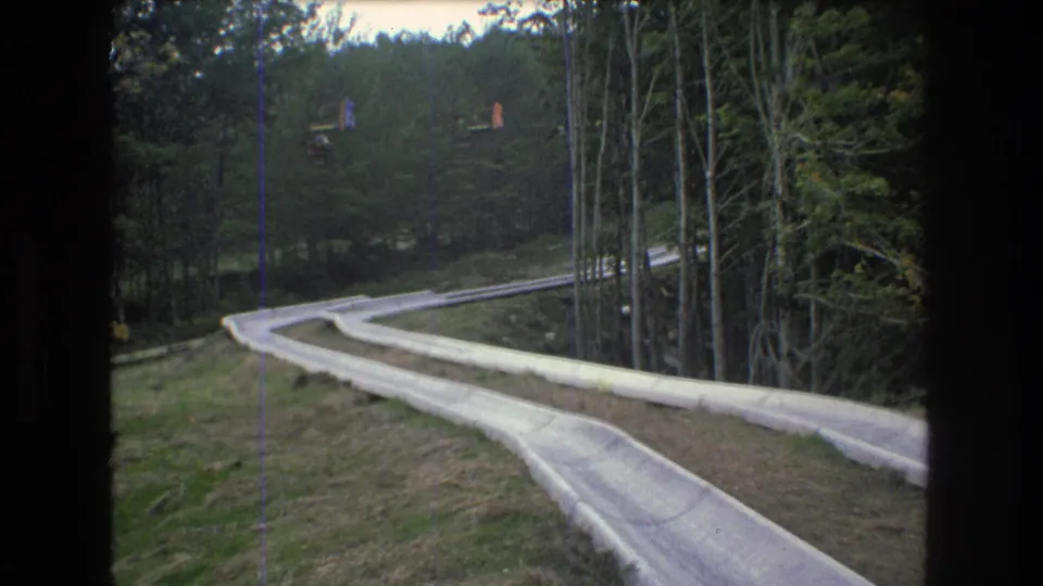 1976 Mt Bromley, Vermont frame looking down the winding concrete slide, with wooded edges, grassy ground, and distant colored lift cars above the treeline.