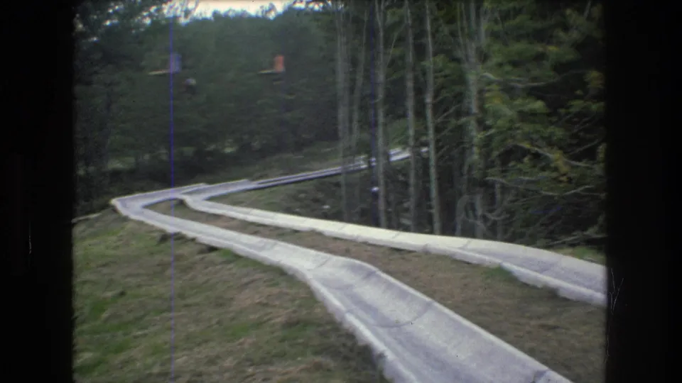 1976 Mt Bromley, Vermont 8mm frame with the slide track running through a wooded resort hillside and faint lift cars or chairs visible above the darker treeline.