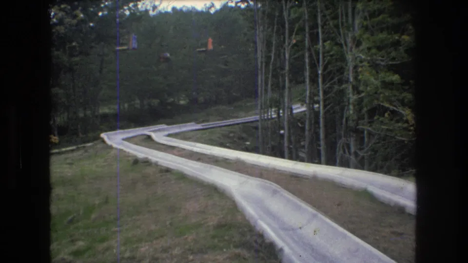 1976 Mt Bromley, Vermont frame showing the pale concrete water slide descending through a forested hillside, with small colorful lift cars visible beyond the trees.
