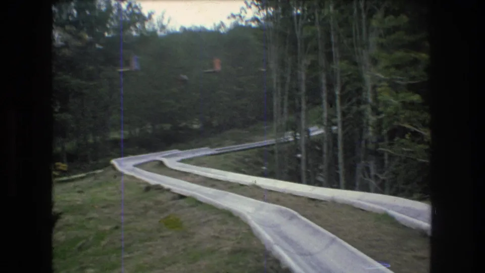 1976 Mt. Bromley, Vermont 8mm frame showing a pale alpine slide track bending downhill through grass and dense trees.