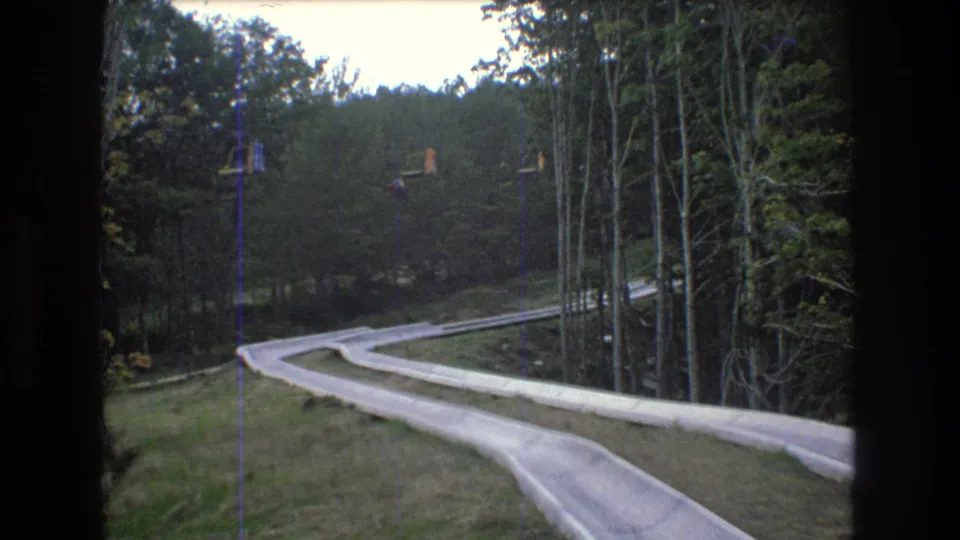 1976 Mt. Bromley, Vermont frame with two pale slide lanes crossing a grassy wooded slope and small distant chairlift shapes above the hill.