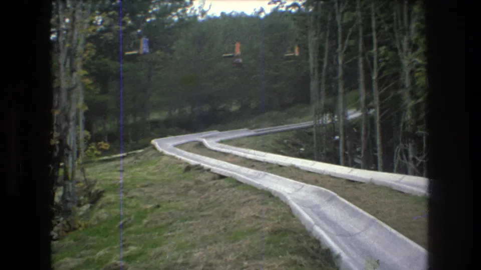 1976 Mt. Bromley, Vermont 8mm still showing the alpine slide track sweeping around a bend beside tall forest trees and a grassy bank.