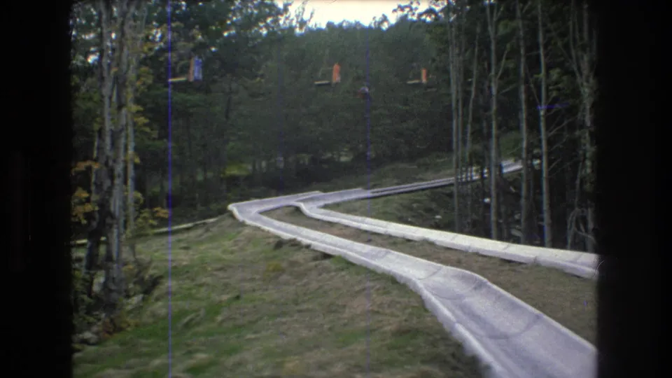 1976 Mt. Bromley, Vermont frame showing a winding alpine slide track disappearing through a wooded mountain slope with lift chairs faintly visible uphill.