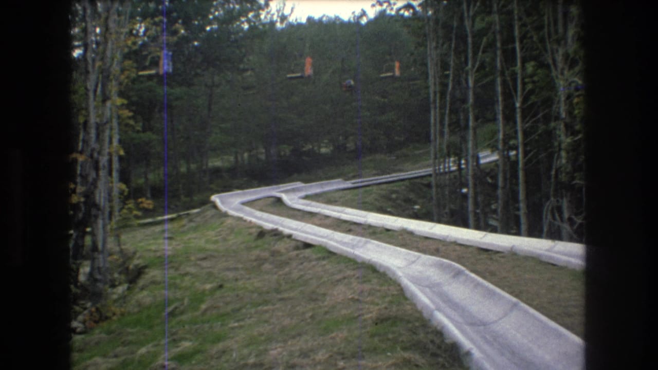 1976 8mm home-movie frame from Mt Bromley, Vermont showing a pale winding outdoor slide trough descending through grass and trees under an overcast sky.
