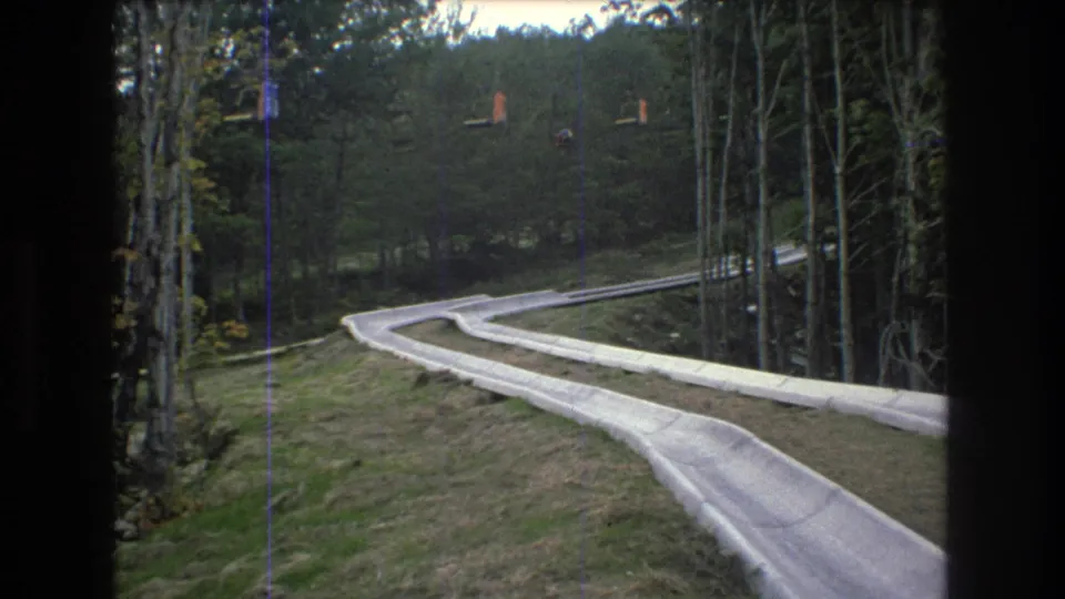 1976 8mm home-movie frame from Mt Bromley, Vermont showing a pale winding outdoor slide trough descending through grass and trees under an overcast sky.