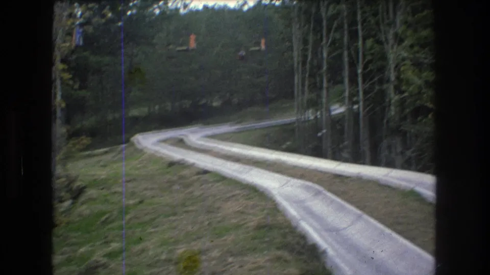 Low-angle 1976 Mt Bromley view of the outdoor slide curving along a grassy slope with forested edges and visible 8mm film wear.