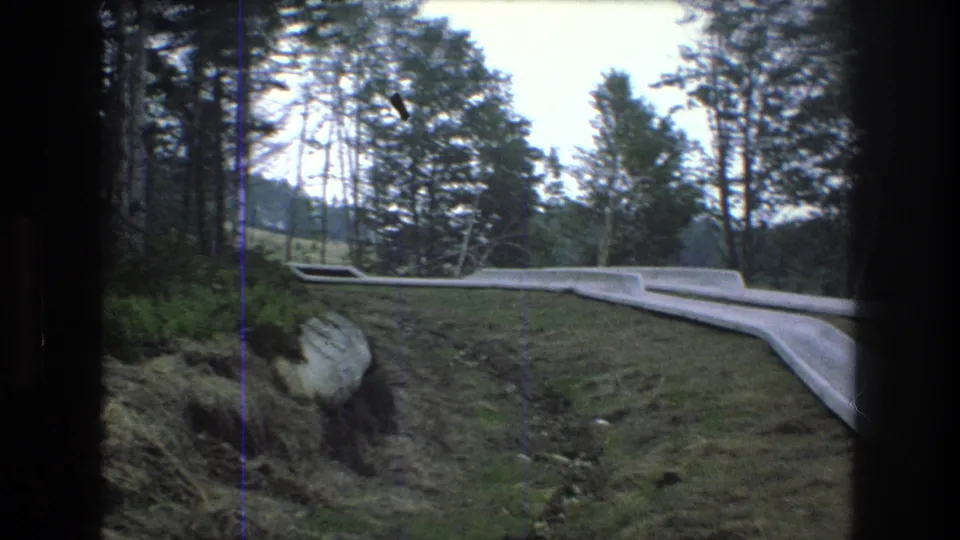 1976 8mm frame showing part of the Mt Bromley outdoor slide beside exposed rocks, grassy ground, and tall trees in a wooded recreation area.