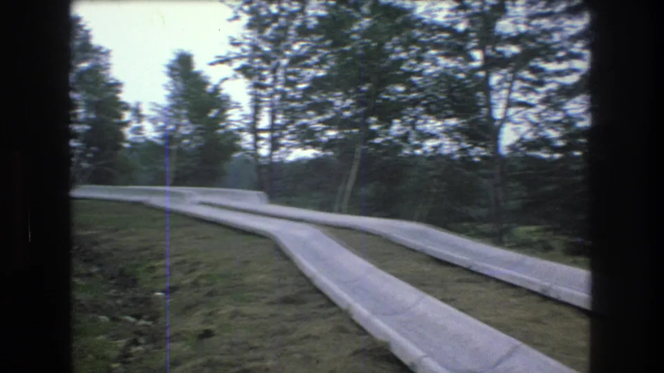 Wide 1976 Mt Bromley home-movie view of the outdoor slide track crossing a grassy clearing with a line of trees in the background.