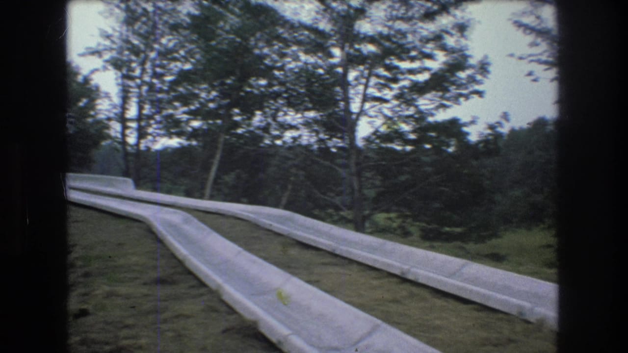 1976 Mt Bromley, Vermont home-movie frame showing a pale concrete path or roadway curving through a wooded hillside with tall trees and grassy edges.
