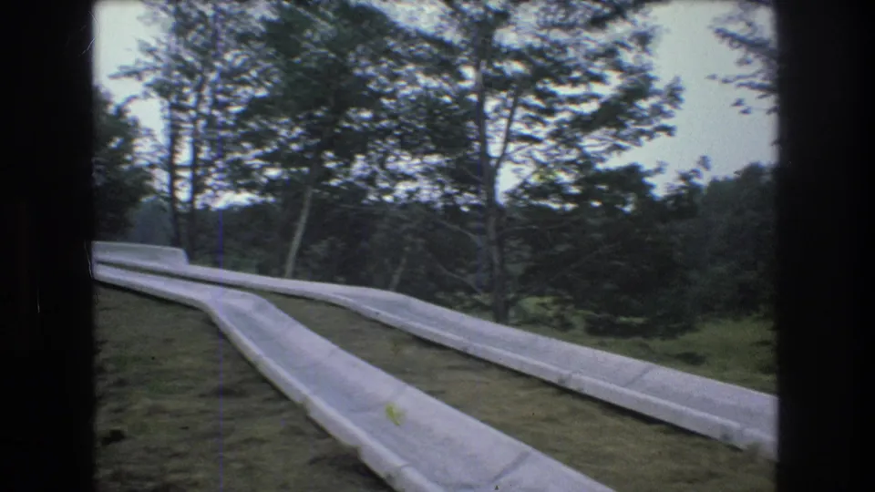 1976 Mt Bromley, Vermont home-movie frame showing a pale concrete path or roadway curving through a wooded hillside with tall trees and grassy edges.