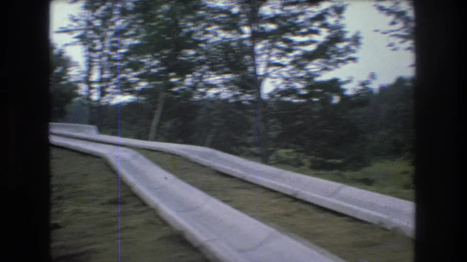 1976 Mt Bromley, Vermont road view with a curving paved lane, pale concrete roadside barrier, grassy shoulder, and trees beyond.