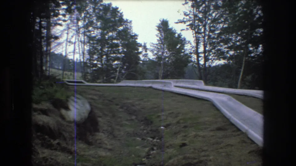 1976 8mm Vermont frame showing the roadside barrier crossing a grassy embankment with rocks, trees, and wooded hills in the background.