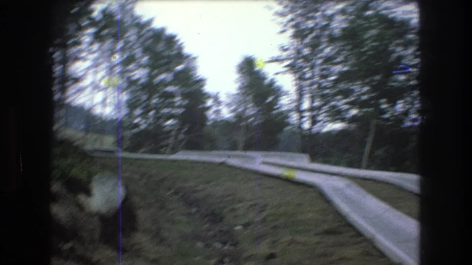 Moving 1976 Mt Bromley, Vermont road shot with blurred roadside trees, a pale concrete barrier, grassy bank, and window framing at the edge.