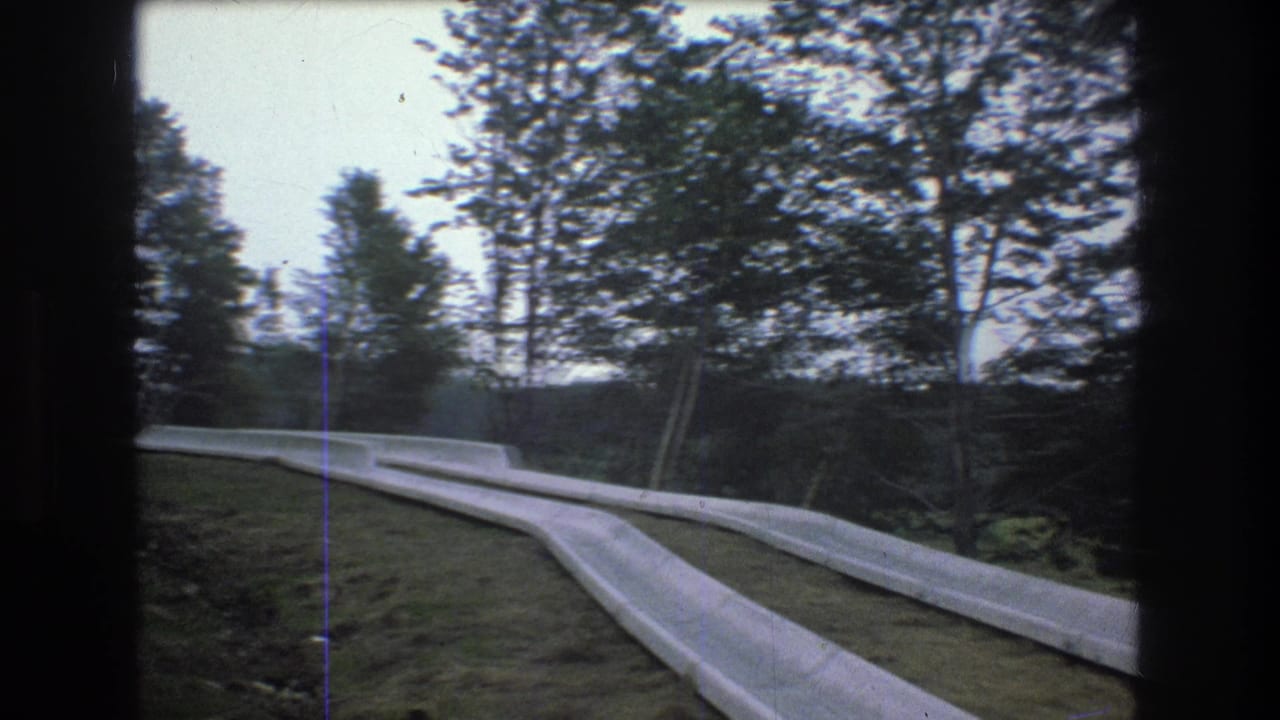 1976 Mt Bromley, Vermont frame with parallel concrete water-slide channels crossing a grassy hillside, backed by dense trees and soft gray sky.