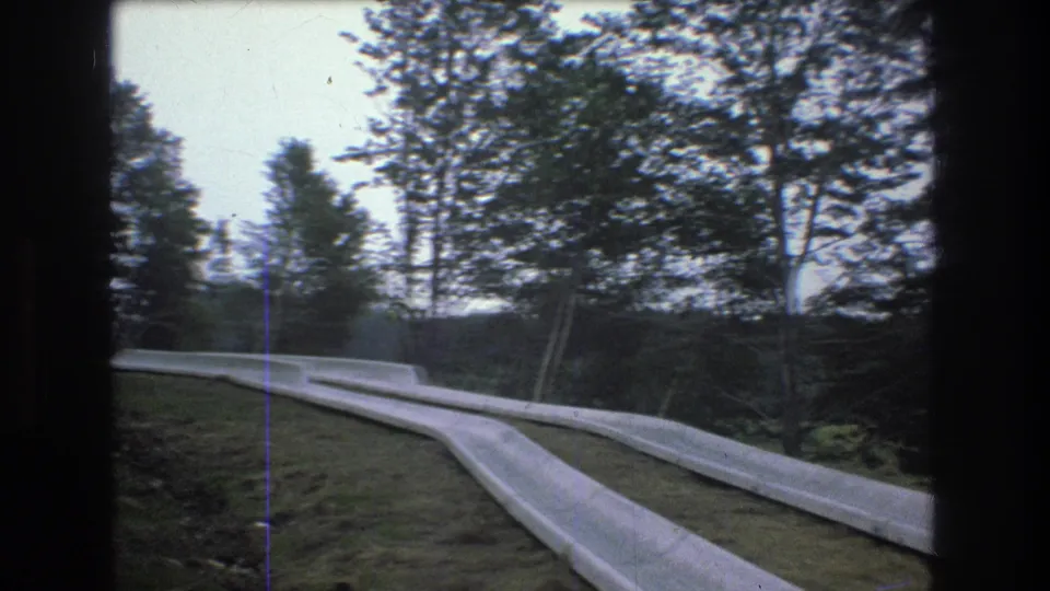 1976 Mt Bromley, Vermont frame with parallel concrete water-slide channels crossing a grassy hillside, backed by dense trees and soft gray sky.