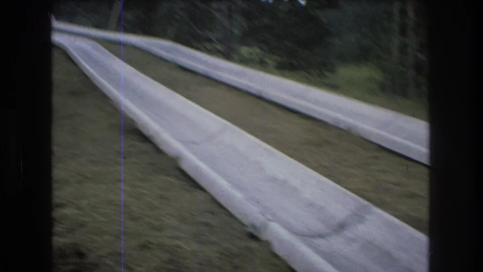 Close 8mm view from Mt Bromley in 1976 looking along pale concrete slide lanes, with motion blur, grass, and dark wooded edges around the frame.