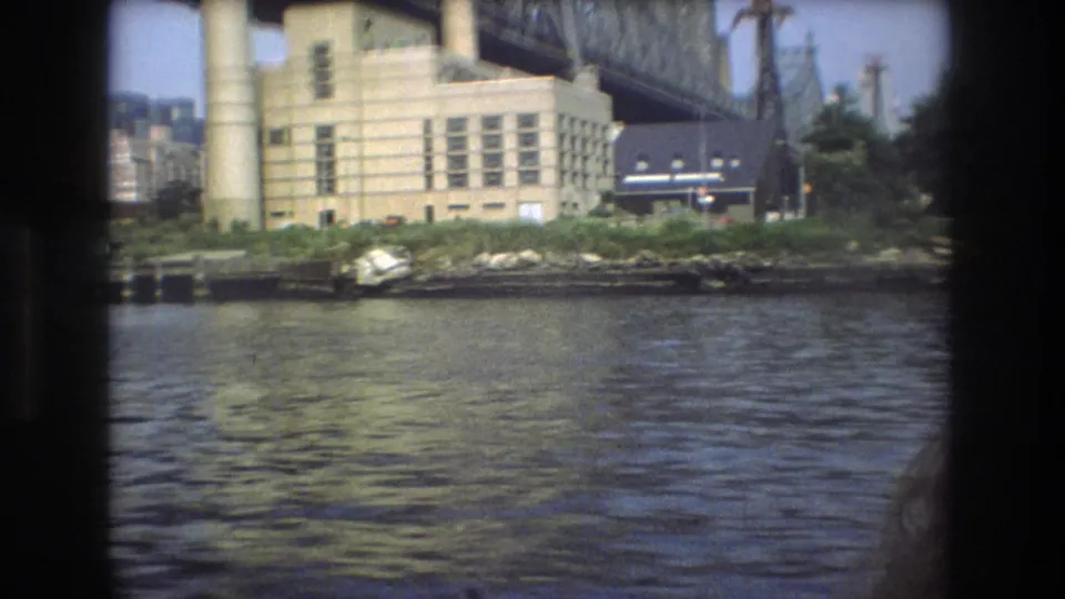 1976 New York City waterfront view from a boat, showing murky river water, a rocky bank, a pale industrial building, smokestacks, and steel bridge structure overhead.