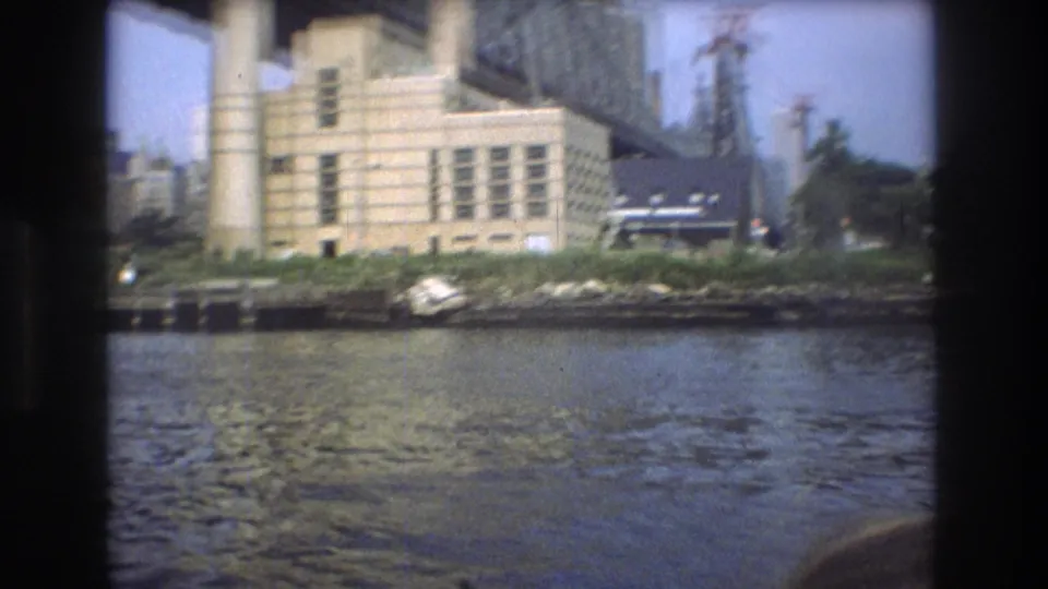 Boat-passenger view of the New York City riverfront in 1976, with a pale industrial building, dark-roofed structure, small boat near the shore, smokestacks, and bridge framing at the top.