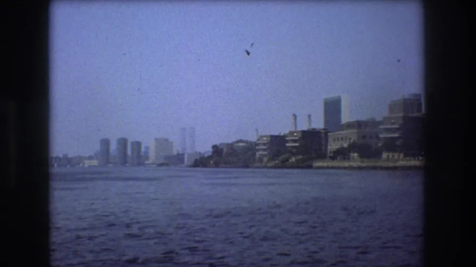 Wide 1976 New York City river view from a moving boat, with hazy distant towers, open water, an industrial shoreline to the right, and soft 8mm color fading.