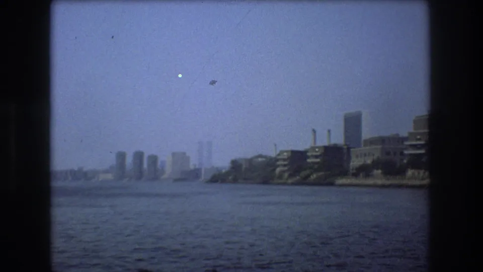 Soft 8mm view of the New York City waterfront in 1976, showing broad river water, distant high-rise towers in haze, and low industrial buildings along the right bank.