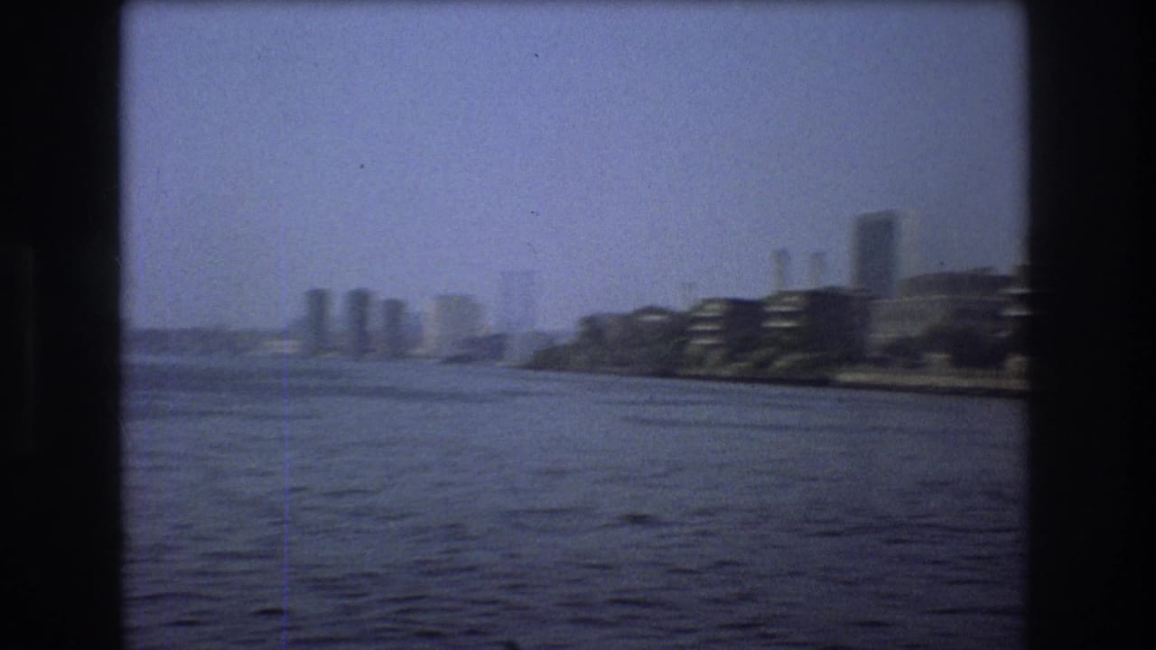 Wide river with distant city skyline and waterfront buildings under a hazy blue sky.
