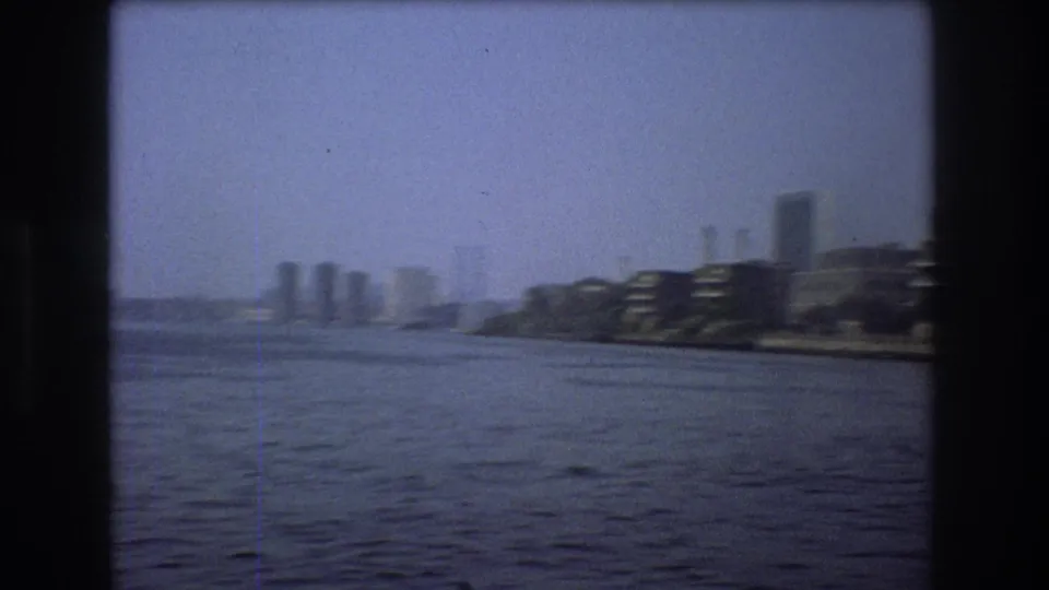 1976 New York City home-movie frame showing a wide river, distant skyline, and low waterfront buildings seen through dark film borders.
