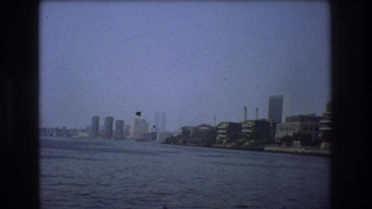 1976 New York City frame with a broad river foreground, hazy skyline in the distance, and waterfront buildings along the right shoreline.