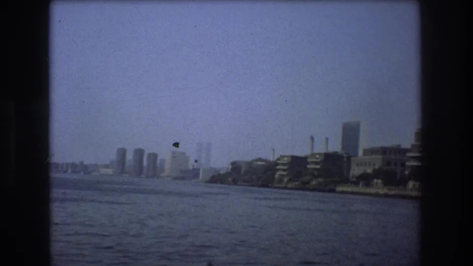 1976 New York City frame with a broad river foreground, hazy skyline in the distance, and waterfront buildings along the right shoreline.