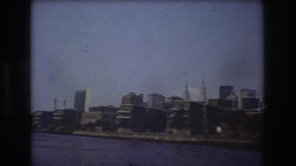 1976 New York City home-movie frame showing a closer waterfront section with low buildings, taller towers behind them, and river water in the foreground.